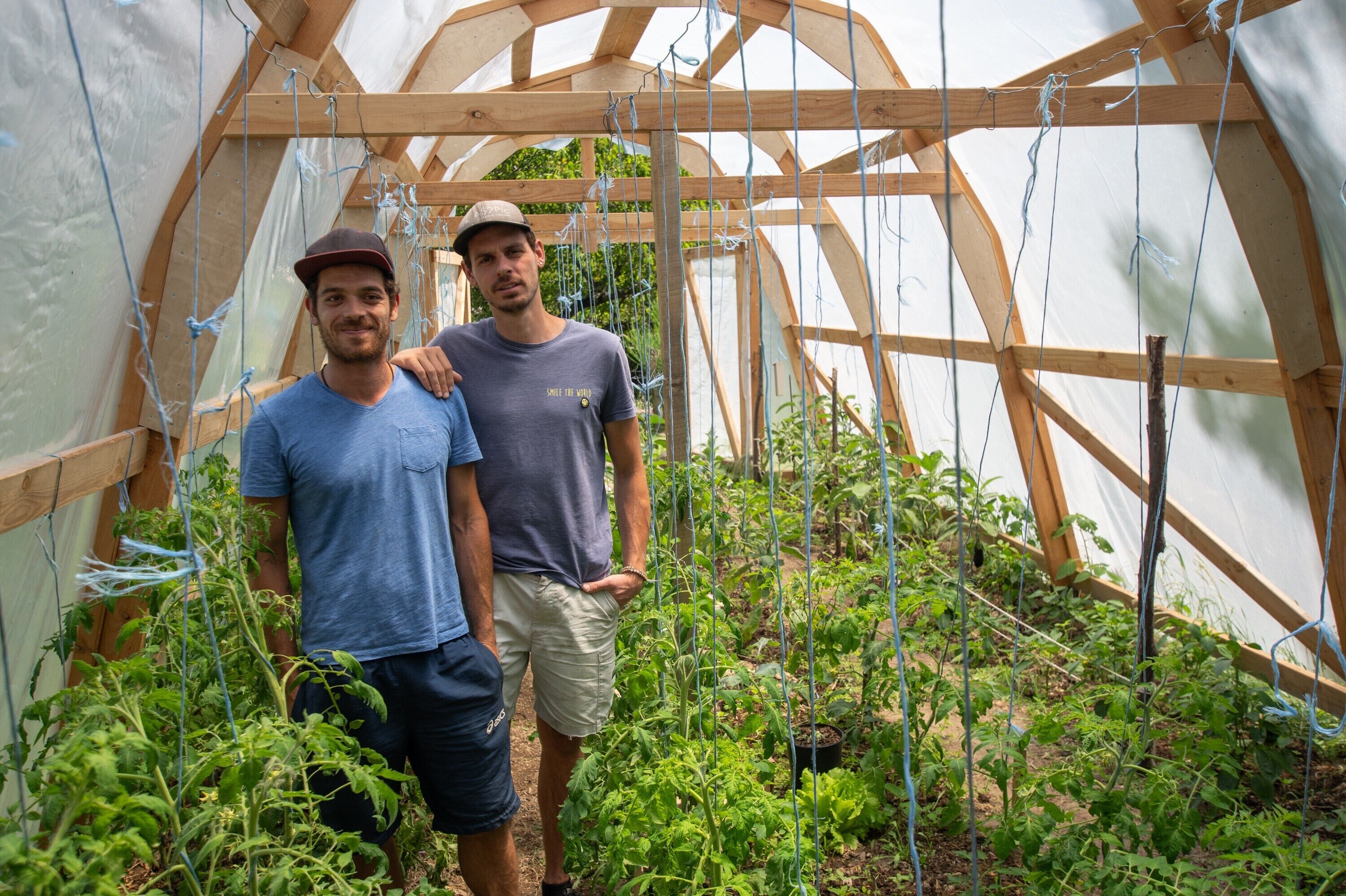 Dos personas de pie en un invernadero con estructura de madera y plantas trepadoras.