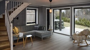 Ett modernt vardagsrum med en grå hörnsoffa, ett soffbord i glas och en gungstol i trä. En trappa med träräcken leder upp till en övre våning.