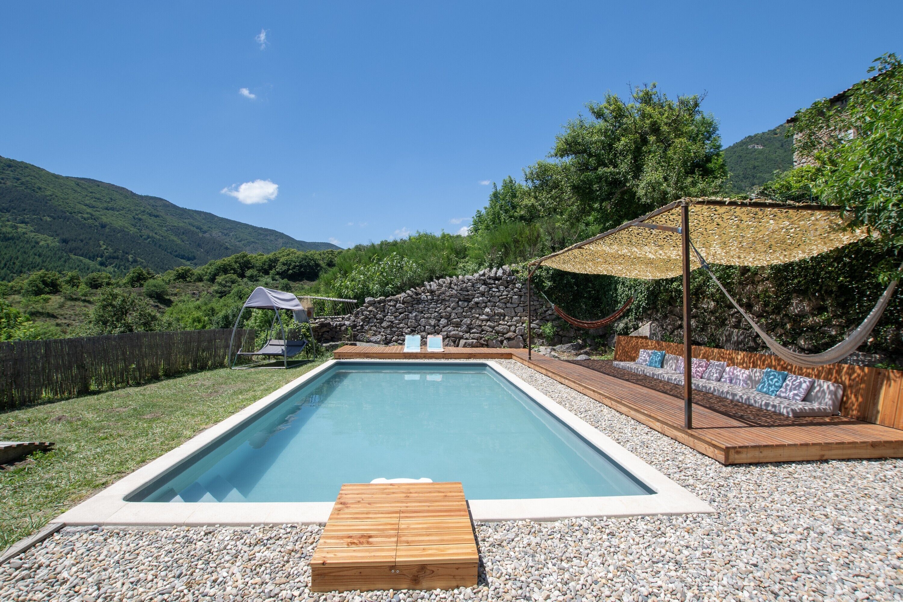Une piscine rectangulaire avec une terrasse en bois, un mur en pierre et un hamac.