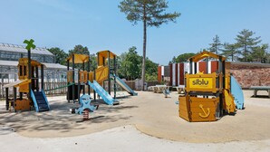 Ein Spielplatz mit Klettergerüsten und Rutschen, einem Picknicktisch und einem Backsteinhaus im Hintergrund.