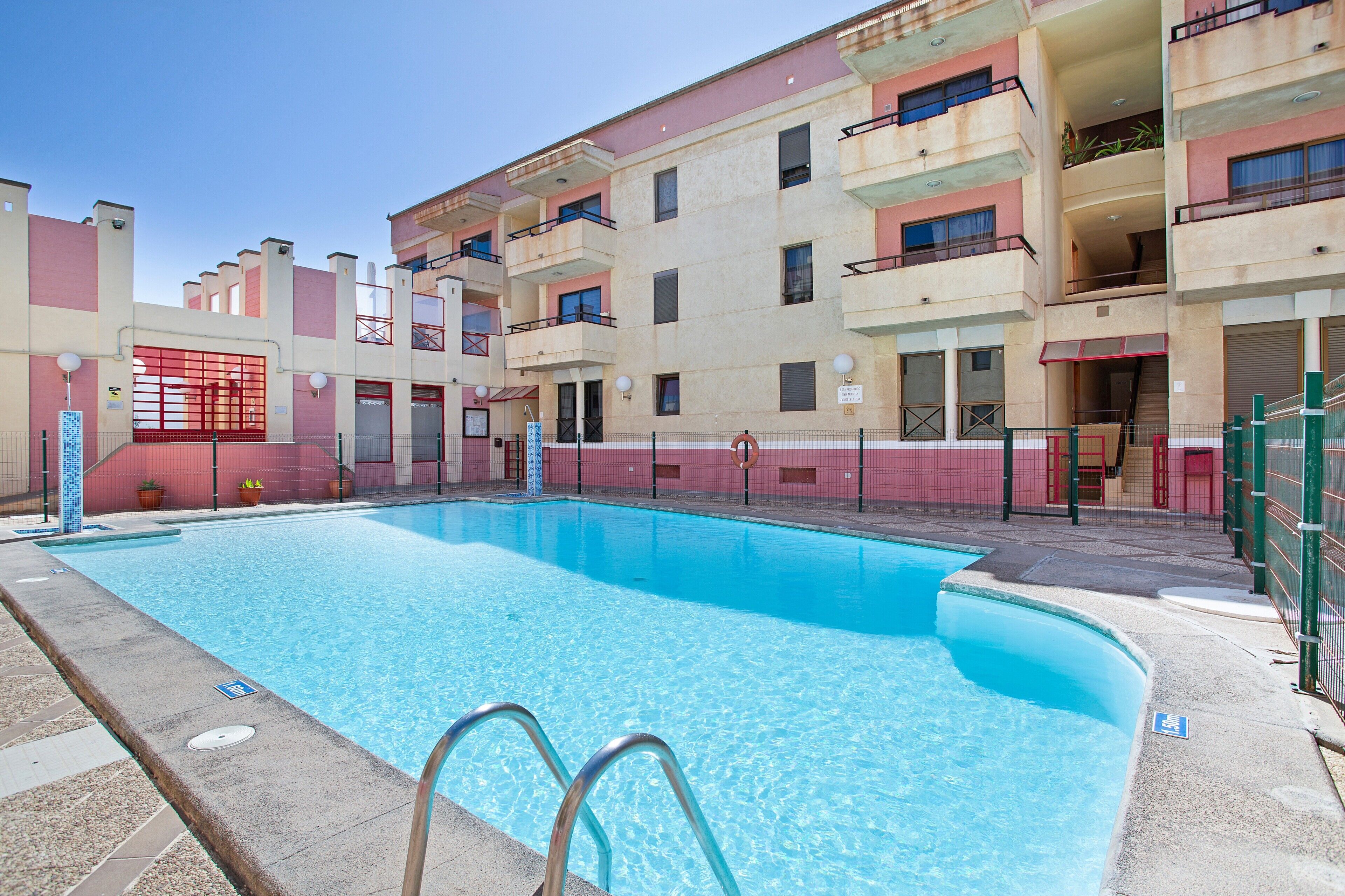 A swimming pool with a metal ladder, surrounded by a multi-story building with balconies and a red and beige color scheme.