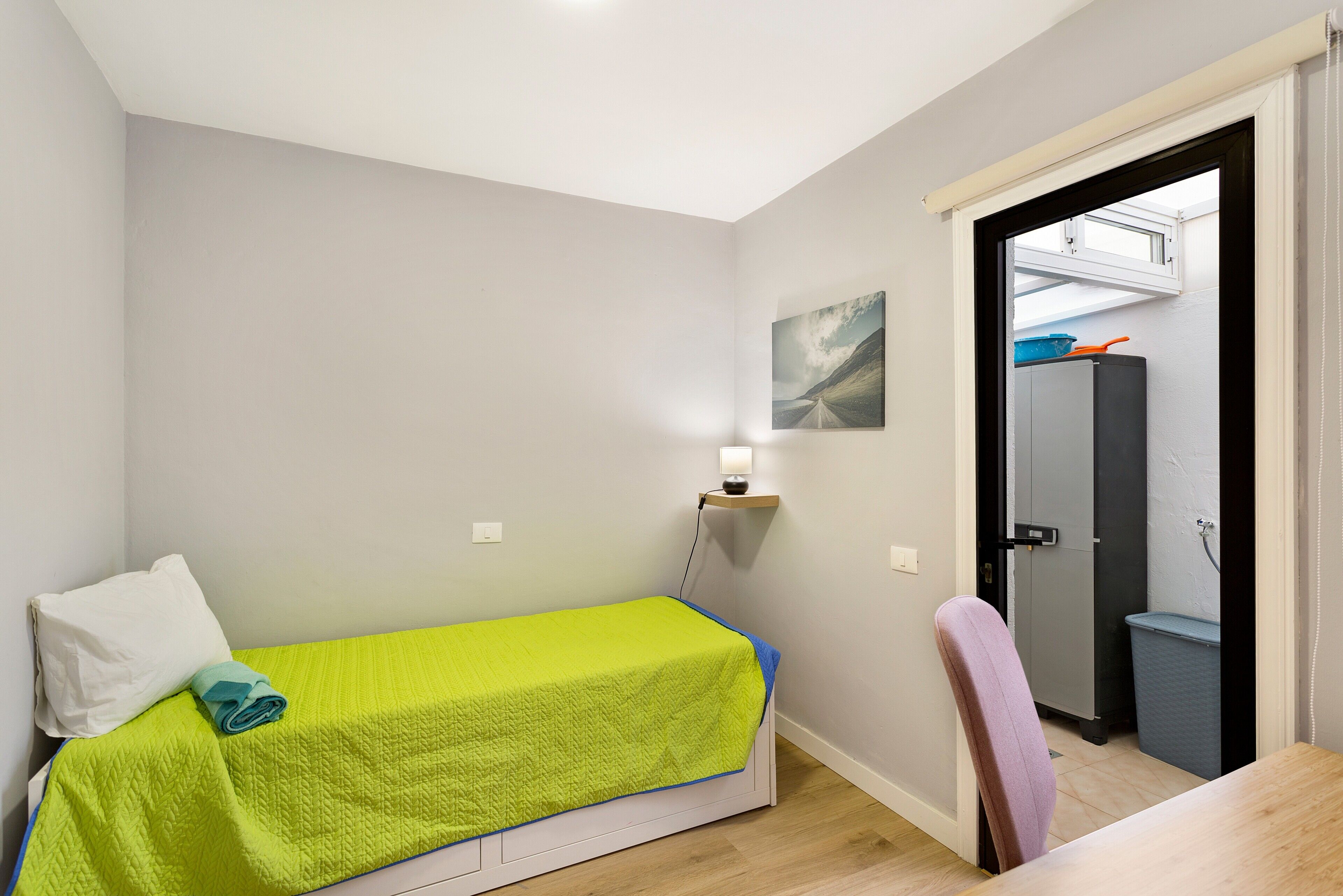 Una habitación pequeña con una cama individual, colcha verde, almohada blanca, una silla, un escritorio, una lámpara de pared y una puerta que da a un baño.