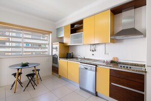 Une cuisine avec des meubles de couleur jaune, un lave-vaisselle, une cuisinière et une fenêtre avec des stores.