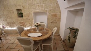 A round wooden table with a white tablecloth, surrounded by white chairs, set against a stone wall with a small window and a potted plant.