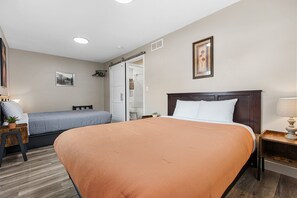 Habitación de hotel con una cama grande, otra más pequeña, un escritorio con lámpara, un cuadro en la pared y una puerta corredera al baño.