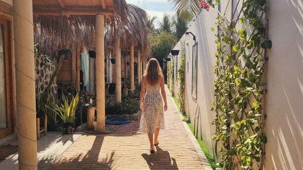 A woman walking down a brick pathway in a covered outdoor area with plants and a thatched roof structure. - Hotel Boutique Puerto (Máncora)
