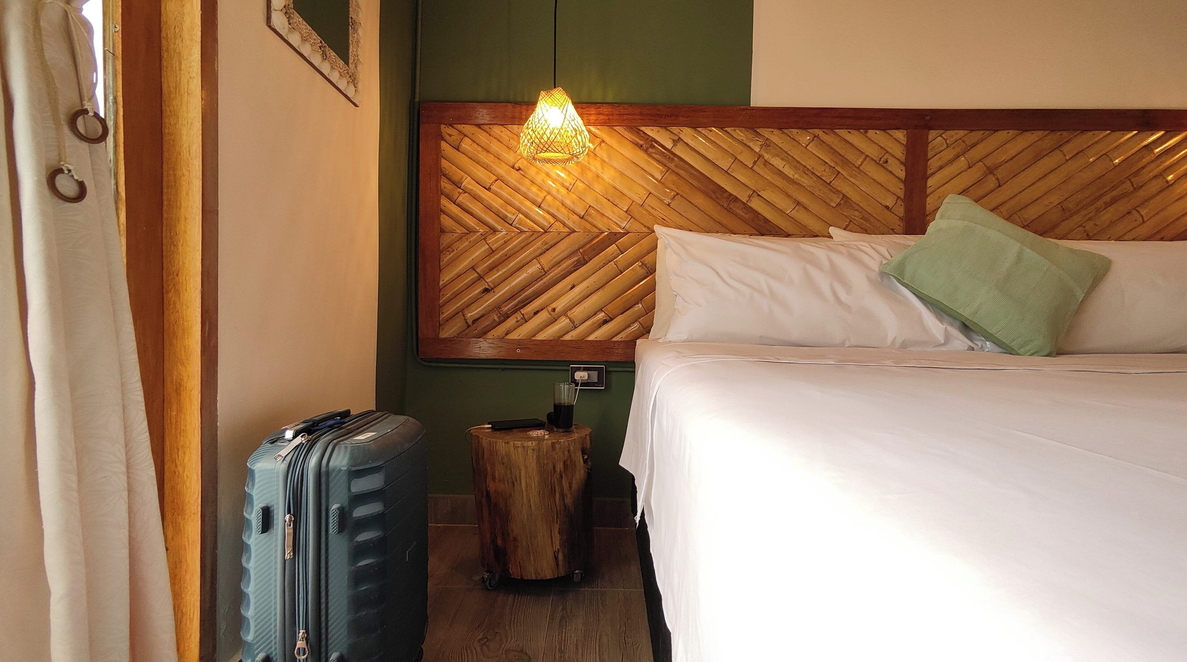 A hotel room with a bed, a nightstand, a lamp, a suitcase, and a wooden headboard.