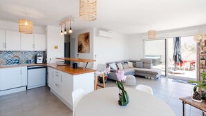 Een moderne keuken met witte kasten, een eethoek met een witte tafel en een woonkamer met een grijze bank.