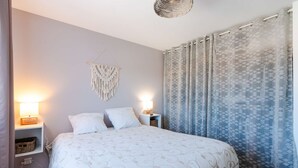 Een net opgemaakt bed met een bedsprei met dessin, twee bedlampjes, een macramé decoratie aan de muur en een gordijn met patroon.