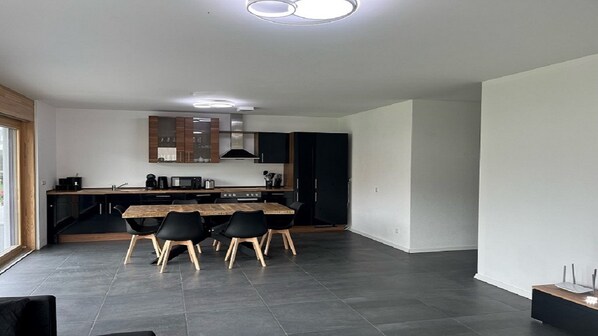 A modern kitchen with a dining area, featuring a large island, black cabinets, and a wooden table with chairs.