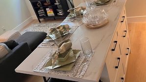 Ein Essbereich mit einem langen Tisch, gedeckt für vier Personen, einer schwarzen Couch, einem Fernseher und einem Bücherregal im Hintergrund.