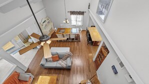 A spacious loft-style living area with a high ceiling, wooden flooring, a central seating area with a sofa and chairs, a dining table, and a ceiling fan. - Enjoy your vacation at the Triple Wave Rider (Virginia Beach)
