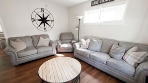Un salón con dos sofás grises, una mesa redonda de centro, una decoración de pared con una brújula y un letrero que dice 'LAKE HOUSE'.