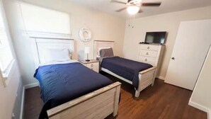 Un dormitorio con dos camas de madera, un ventilador de techo, un televisor sobre un aparador y una puerta.
