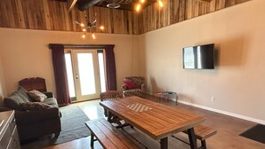 Un salón espacioso con una mesa de madera, bancos, un sofá y un televisor montado en la pared.