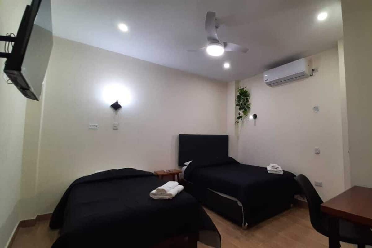 A small, clean room with two single beds, a desk, a chair, a ceiling fan, and an air conditioning unit.