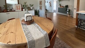 A dining area with a wooden table, chairs, and a patterned table runner. There are potted plants and wall art on shelves.