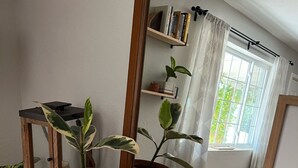 A room with a large mirror reflecting a window with curtains, a bookshelf, and two potted plants on a white dresser.