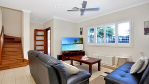 A living room with a sofa, a coffee table, a TV displaying a beach scene, a ceiling fan, and a window with blinds. - Bennett’s Beach Townhouse (Hawks Nest)