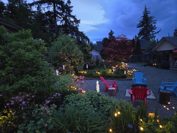 A garden with various plants and trees, outdoor seating with chairs, and decorative lights.