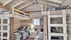 Een rustieke blokhut met houten wanden en een schuin dak. Er staan twee stapelbedden met ruitjes beddengoed en een klein tafeltje met een lamp.