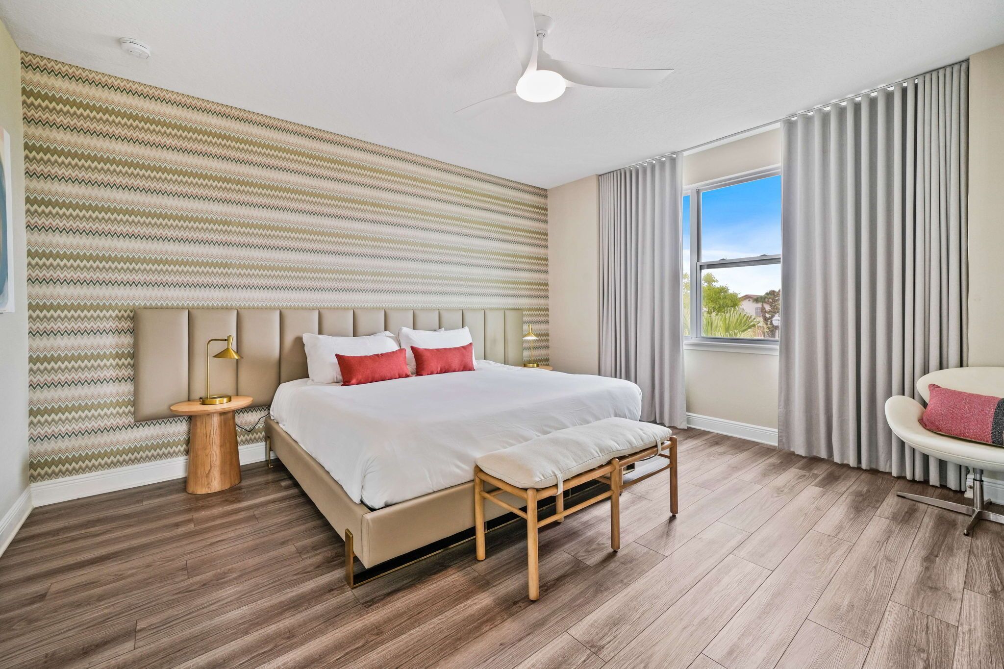 Un dormitorio moderno con una cama grande, una mesita de noche de madera, un ventilador de techo y vistas al exterior a través de una ventana.