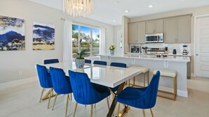 A modern kitchen with white cabinets, a central island, and a dining area with a white table and blue chairs. - Blue Suede Townhome in Reunion Resort (Kissimmee)