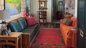 A cozy living room with a stone wall, wooden beams, a red rug, a green sofa, an orange sofa, and a bookshelf. - NEW! Historic Guest Home on Main - Heart of Town (Salado)