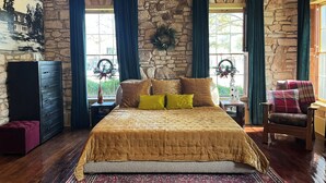 Una habitación con pared de piedra, cama con cubrecama dorado, cómoda de madera y ventana con cortinas.