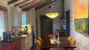A dining area with a wooden table and chairs, a kitchen with modern appliances, and a stone wall with a painting. - NEW! Historic Guest Home on Main - Heart of Town (Salado)