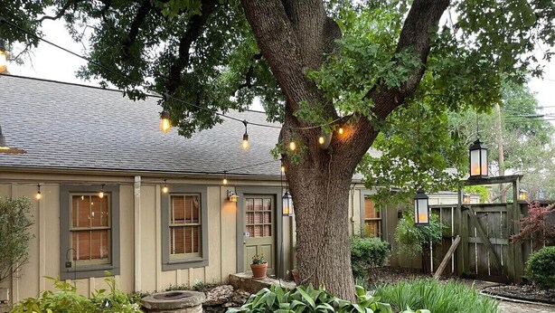 A house with a large tree in the yard, string lights, and a garden with various plants. - NEW! Historic Guest Home on Main - Heart of Town (Salado)