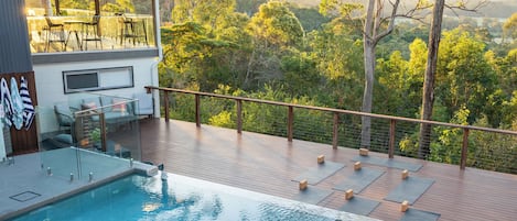 A swimming pool with a wooden deck, lounge chairs, and a view of a forested landscape.