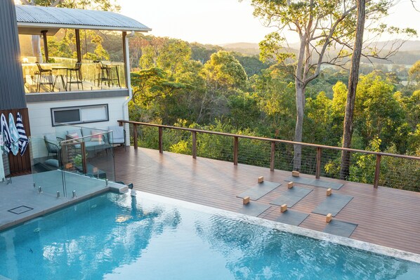 A swimming pool with a wooden deck, lounge chairs, and a view of a forested landscape.