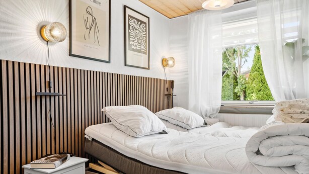 Un dormitorio moderno con cabecera de listones de madera, una cama con ropa de cama blanca, una mesita de noche con un libro y cuadros en la pared.