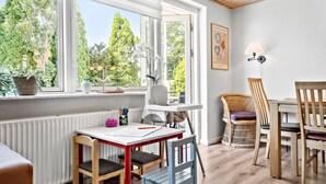 Una habitación con una mesa, sillas, un radiador, una ventana que da a árboles y una lámpara fijada a la pared.