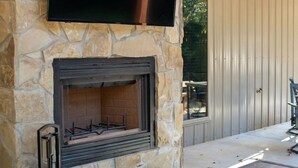 A stone fireplace with a flat-screen TV mounted above it, a metal table, and a patio area with a view of a building in the background. - Cozy Lodge at Ward Creek Ranch close to interstate and wedding venues.  (Simms)