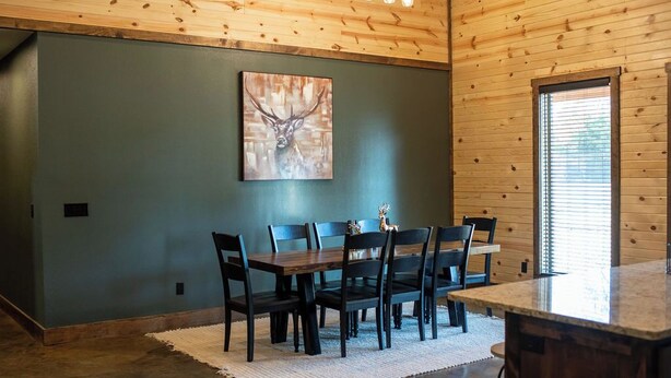 A dining area with a wooden table and chairs, a painting of a deer head, and a window with blinds. - Cozy Lodge at Ward Creek Ranch close to interstate and wedding venues.  (Simms)