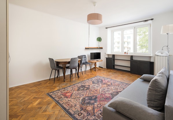 A modern living room with a grey sofa, a patterned rug, a dining area with chairs and a table, and a kitchen with a TV and plant.