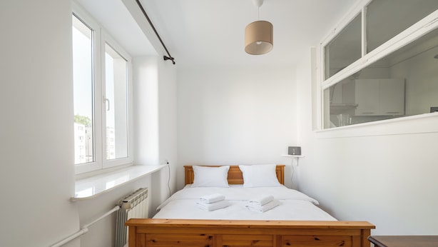 Une chambre à coucher avec un lit en bois, du linge de lit blanc, une fenêtre et un radiateur.