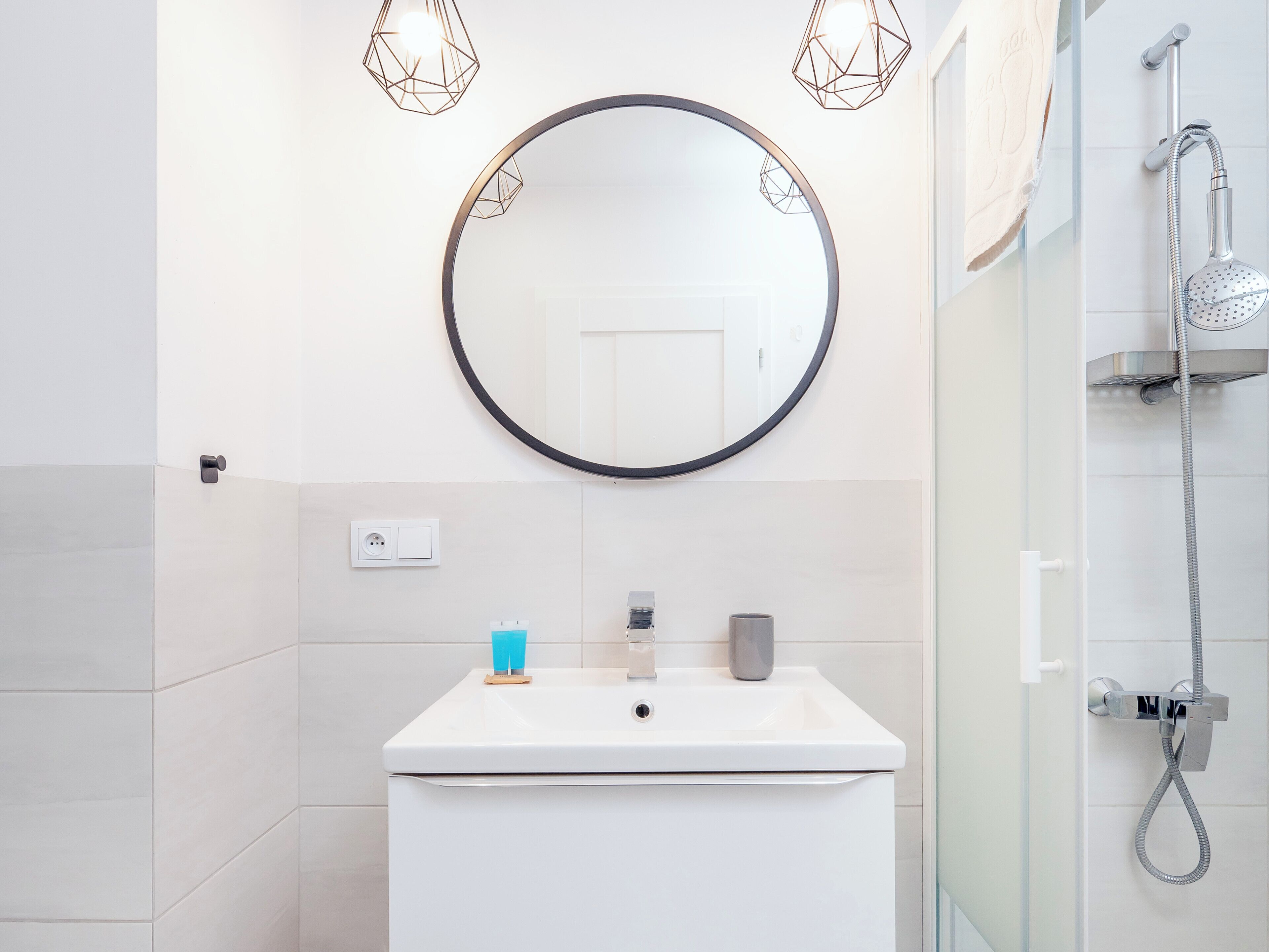 A modern bathroom with a round mirror, a white sink, a handheld showerhead, and two geometric pendant lights.