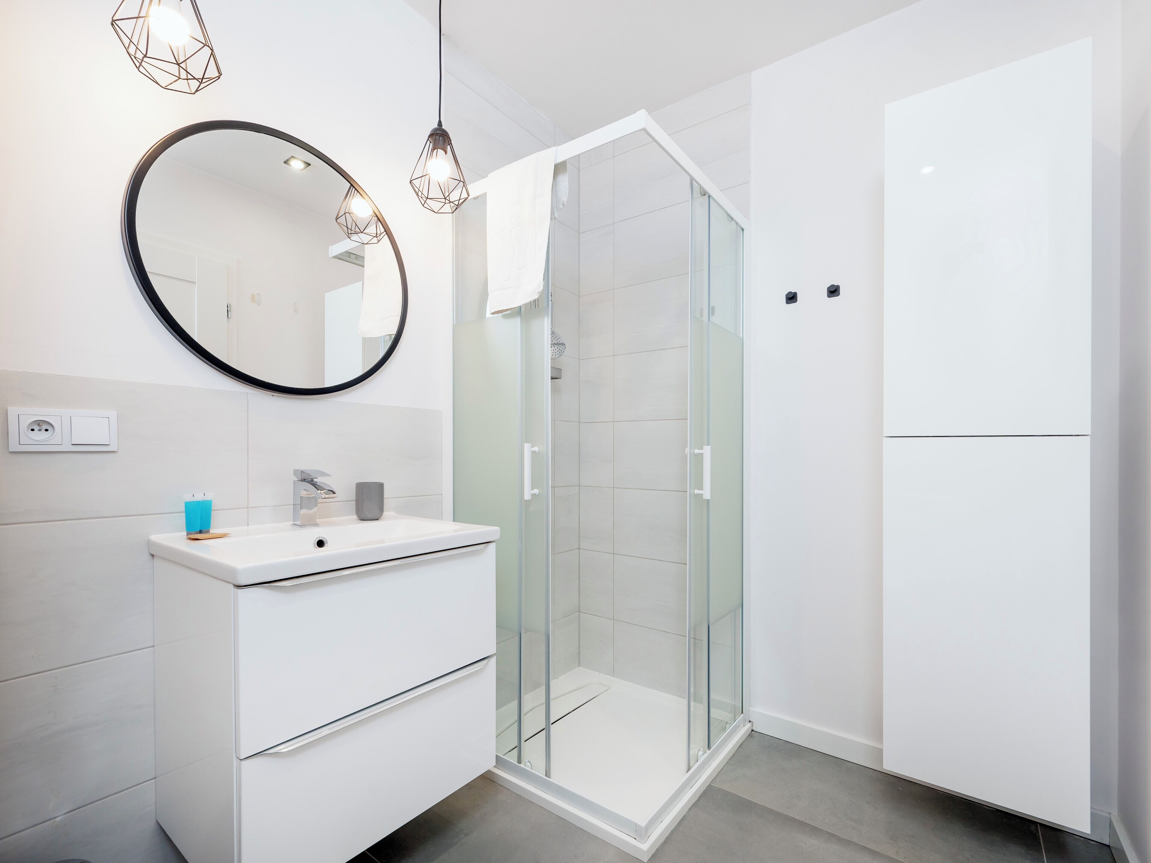Une salle de bain moderne avec une douche à l’italienne en verre, un meuble-lavabo blanc avec un miroir et des luminaires suspendus.