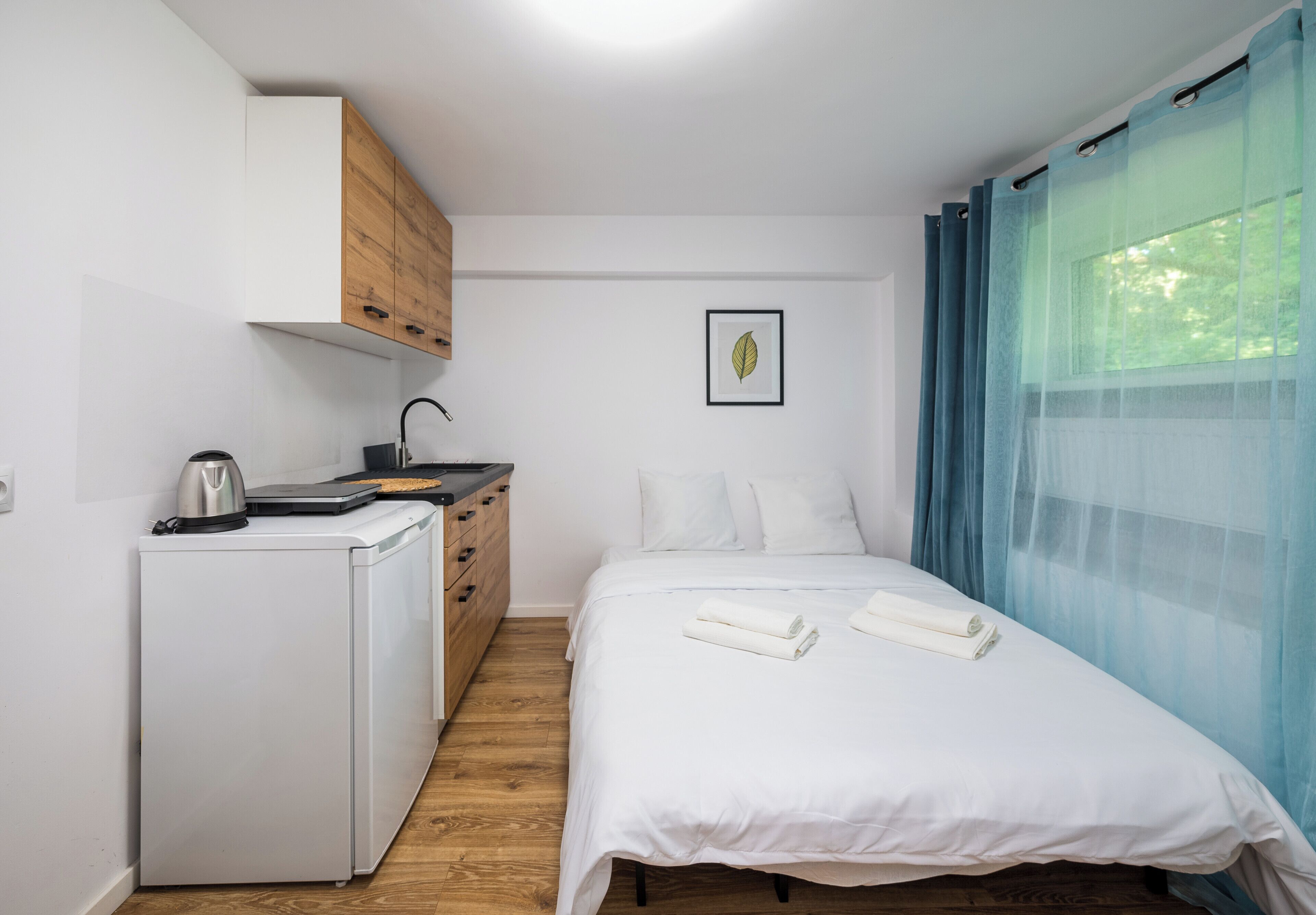 Une chambre à coucher de taille réduite, comprenant un lit, un petit coin cuisine et une fenêtre agrémentée de rideaux légers.