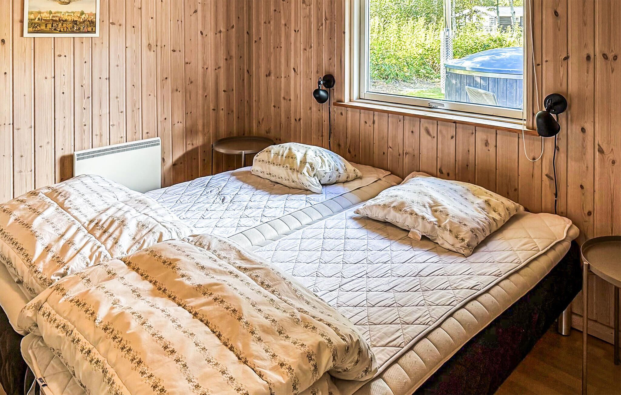 A wooden-paneled room with a bed, a small round table, and a window with a view of greenery and a blue structure outside.