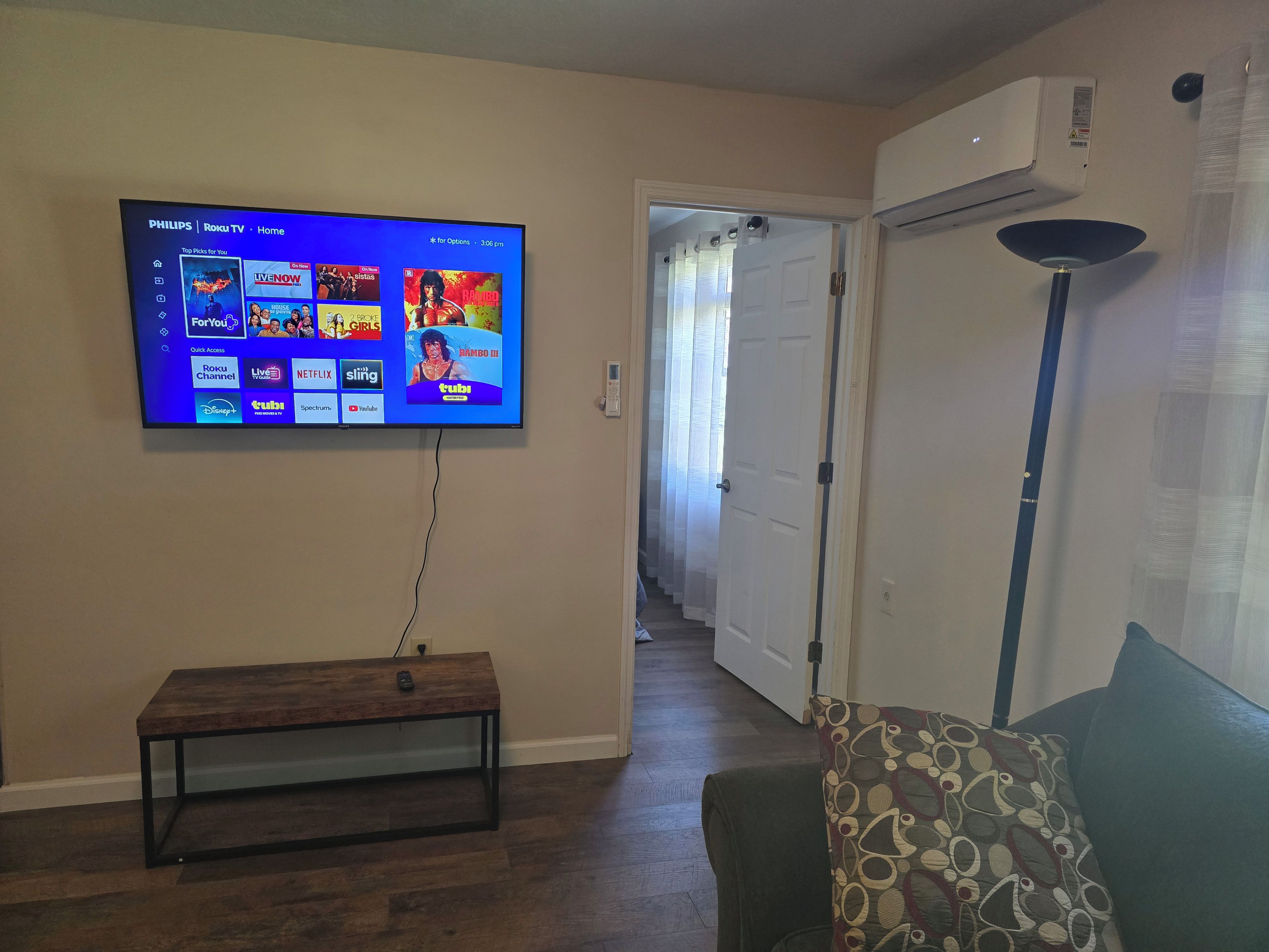 Un salon avec un téléviseur fixé au mur diffusant divers services de streaming, un banc en bois, un canapé avec des coussins à motifs et un climatiseur fixé au mur.