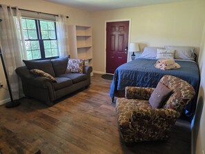 A room with a sofa, a patterned armchair, a bed with a blue bedspread, a red door, and a window with curtains.