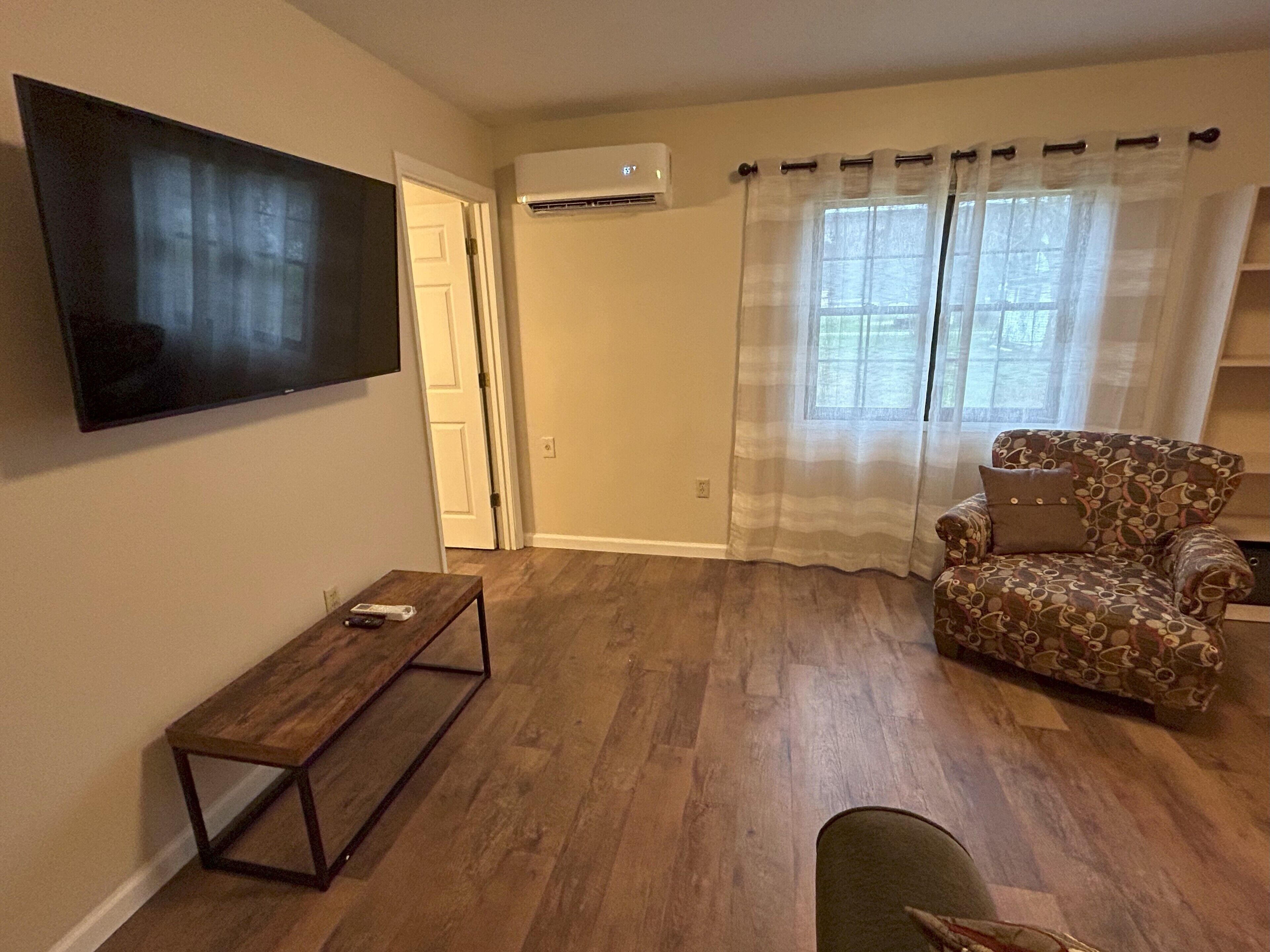 Ein Wohnzimmer mit Holzdielenboden, einem an der Wand befestigten Flachbildfernseher, einem gemusterten Sessel und einem kleinen Holztisch.