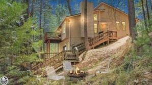 Une cabane en rondins à deux étages, dotée d’une terrasse et d’un foyer extérieur, le tout mis en valeur au premier plan.