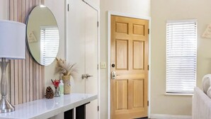 Un couloir avec une porte en bois, une console blanche surmontée d’un miroir et une fenêtre avec des stores.