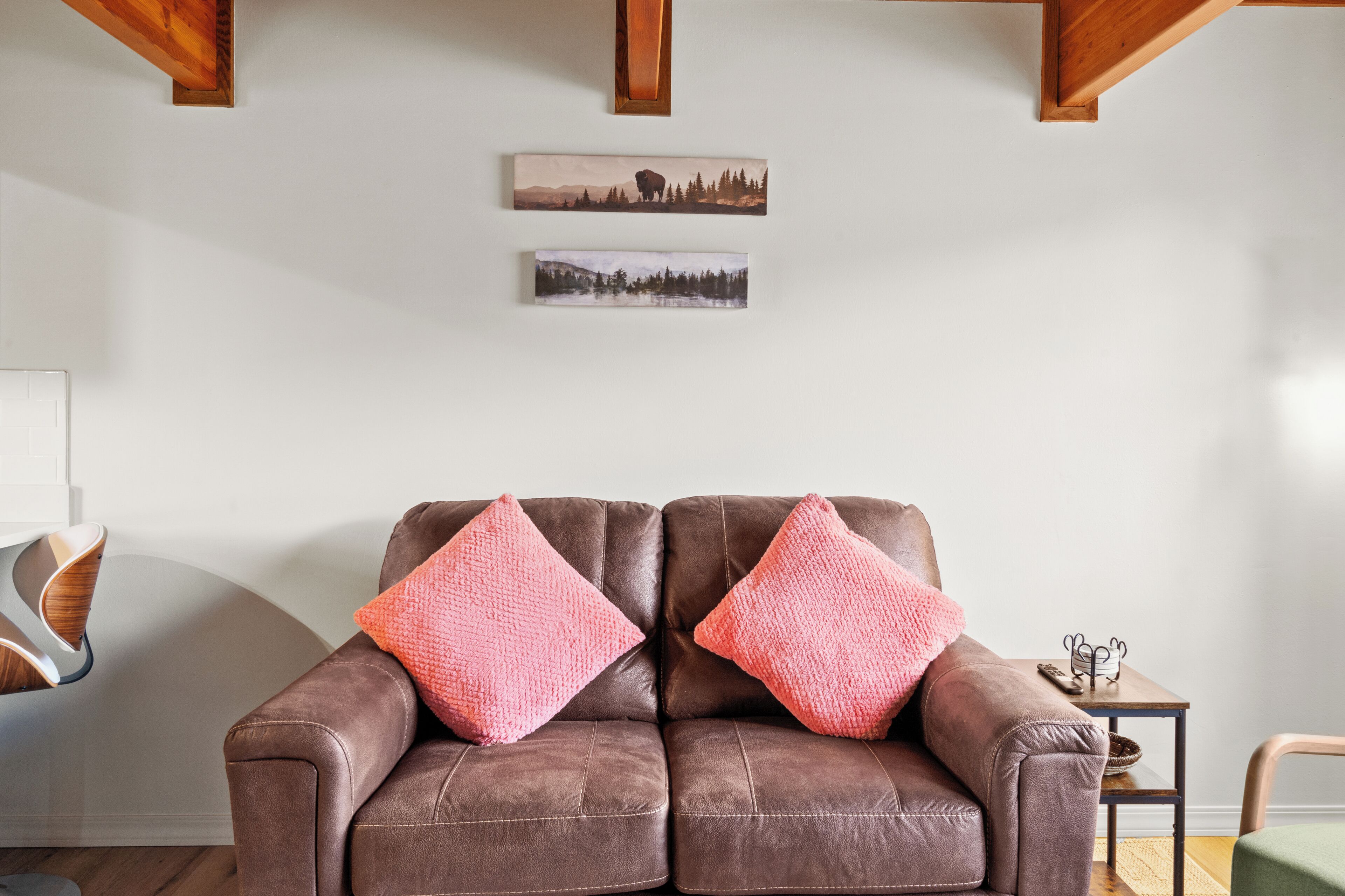 A brown leather sofa with two pink cushions, a small wooden table with a metal object on it, and two framed pictures on the wall.