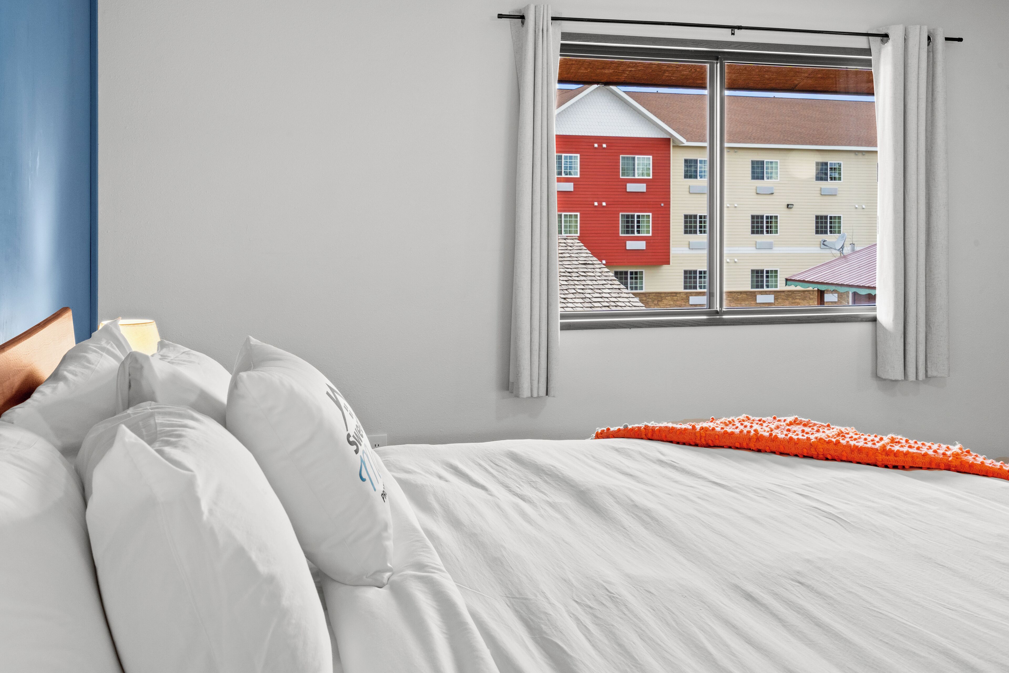 Une chambre à coucher avec un lit, des oreillers, une fenêtre donnant sur des immeubles et un mur bleu.